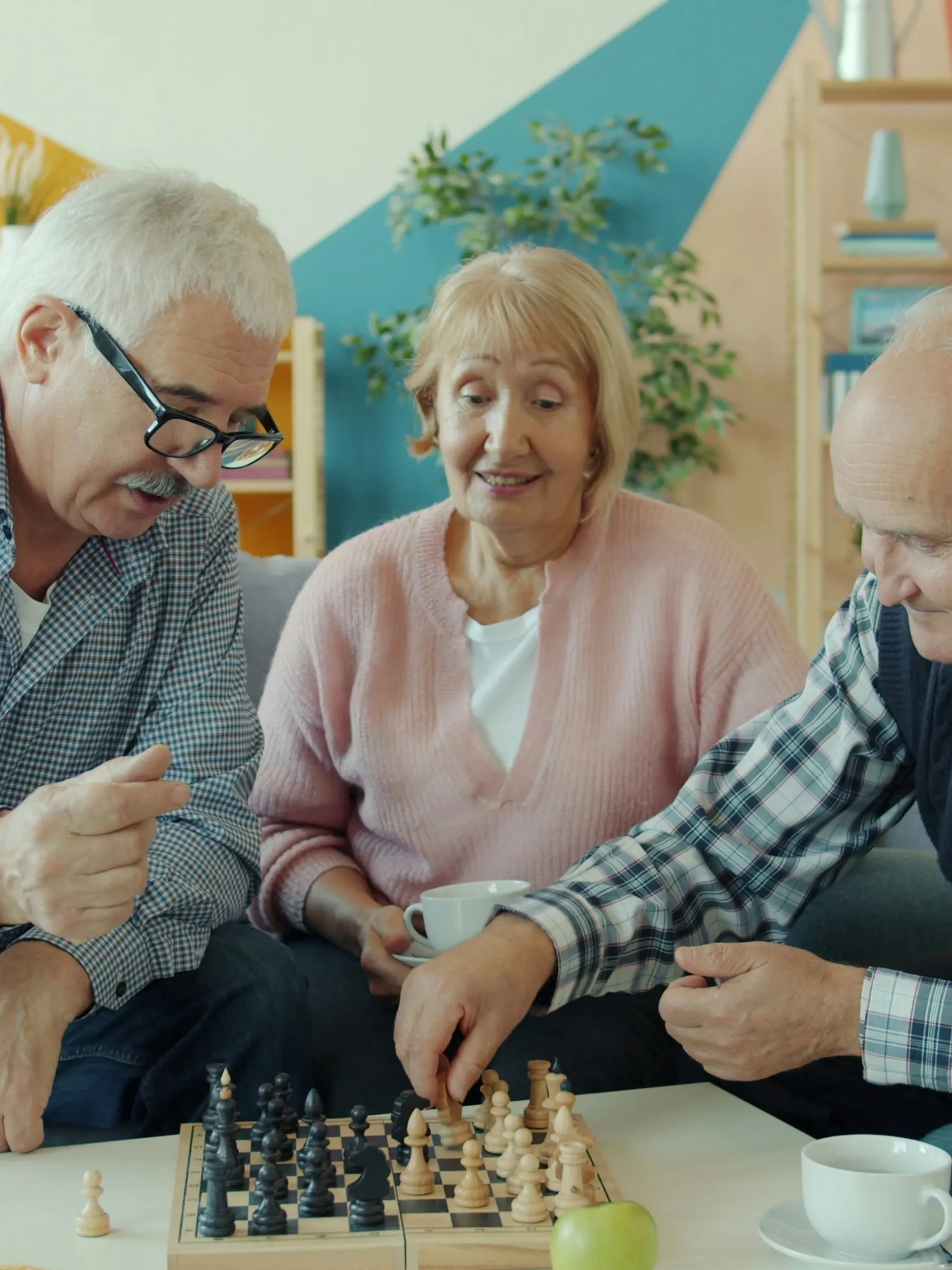 Drei ältere Menschen sitzen gemeinsam im Wohnzimmer, zwei spielen Schach