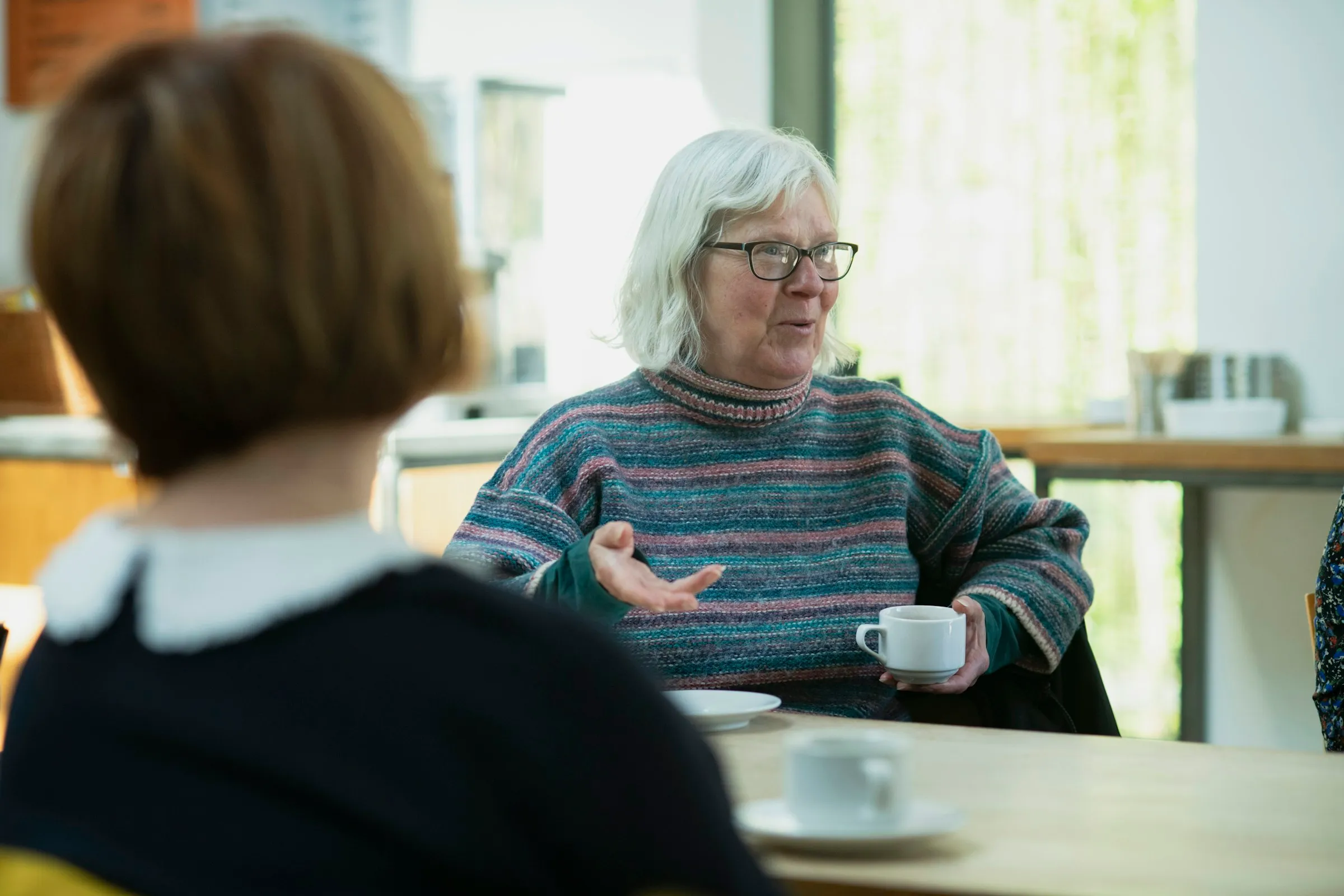 Ältere Frau im Gespräch an einem Tisch, vor ihr eine Tasse